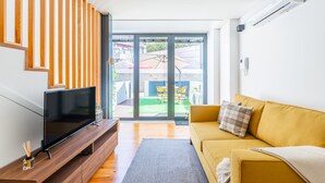 Een moderne woonkamer met een flatscreen televisie op een houten tv-meubel, een gele bank met een geruit kussen, en een glazen schuifdeur die leidt naar een terras buiten met een tafel en stoelen.