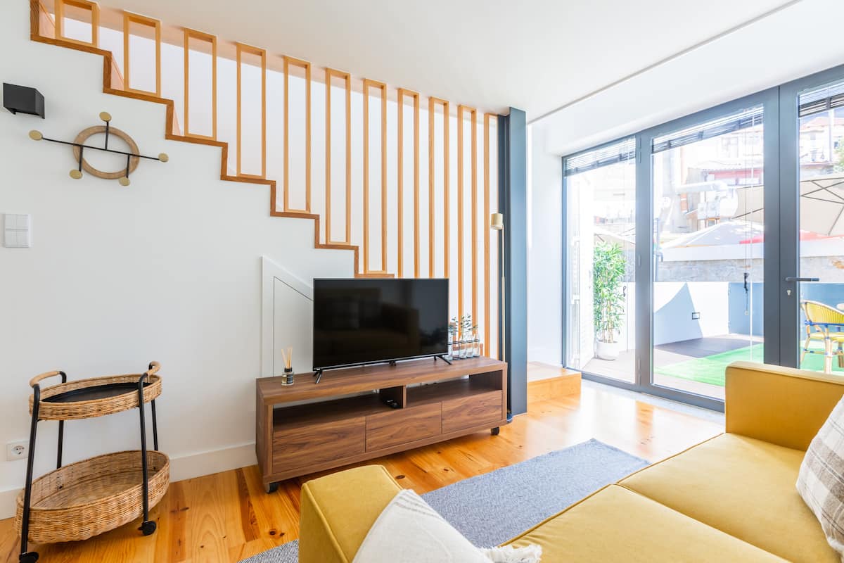 A modern living room with a wooden staircase, a flat-screen TV on a wooden console, a wicker side table, and a sofa.