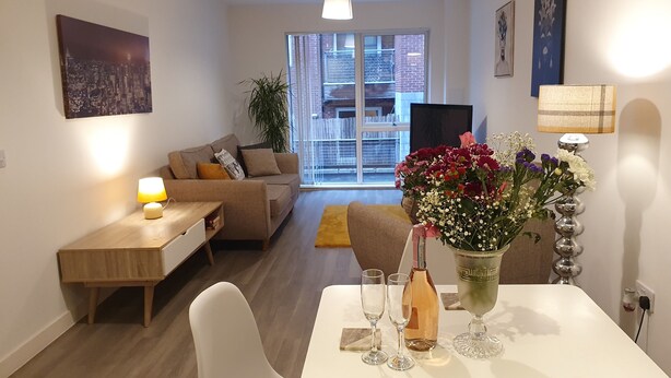 A modern living room with a sofa, a dining table with glasses and a bottle, and a large window with a view of buildings. - Stylish 2-bed Apartment in Manchester City Center (Manchester)