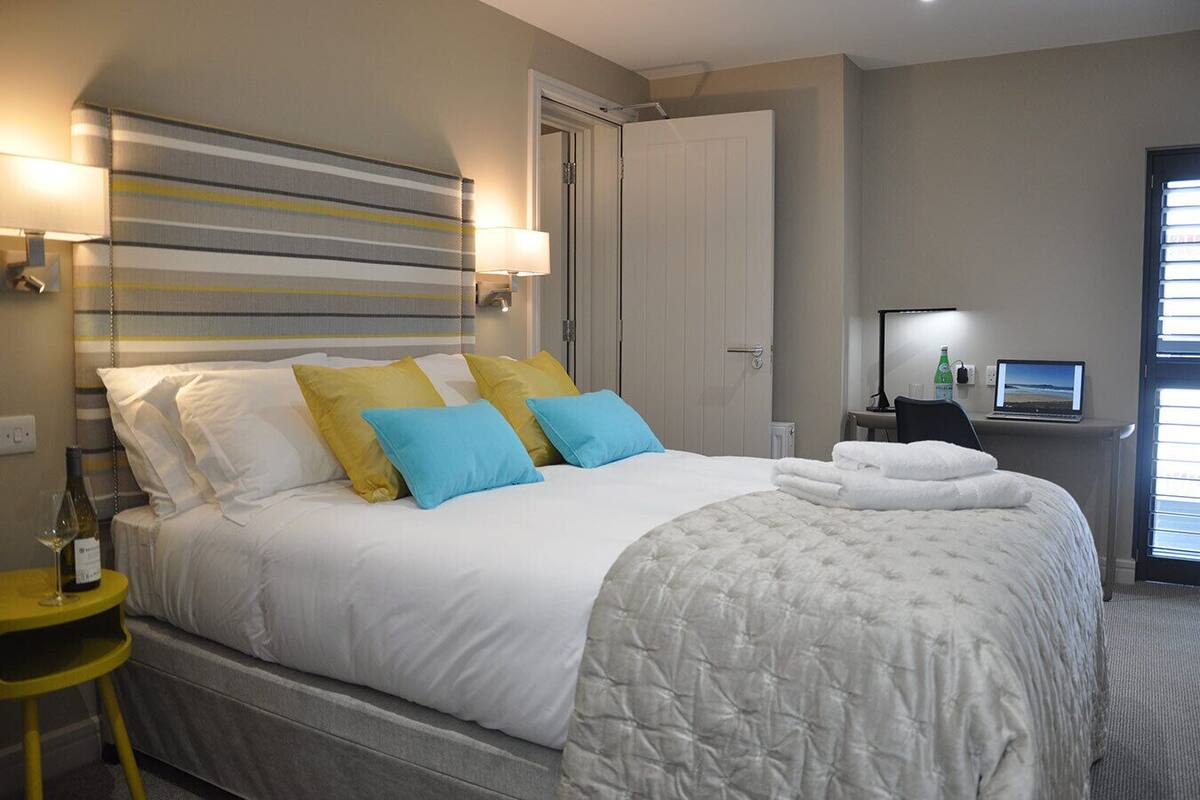 A hotel room with a large bed, a bedside table with a lamp, a laptop, and a view of the outside through a window.