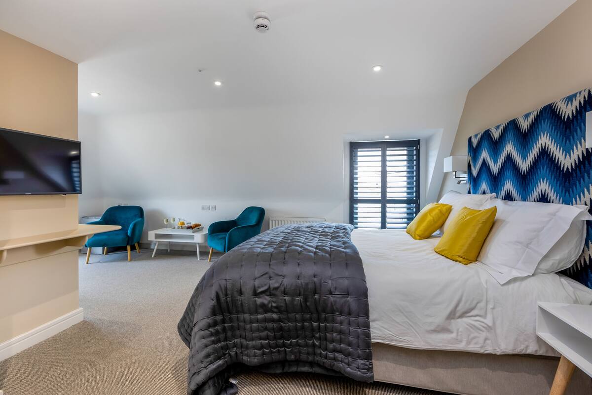 A modern hotel room with a large bed, two blue armchairs, a small white table, and a TV mounted on the wall.
