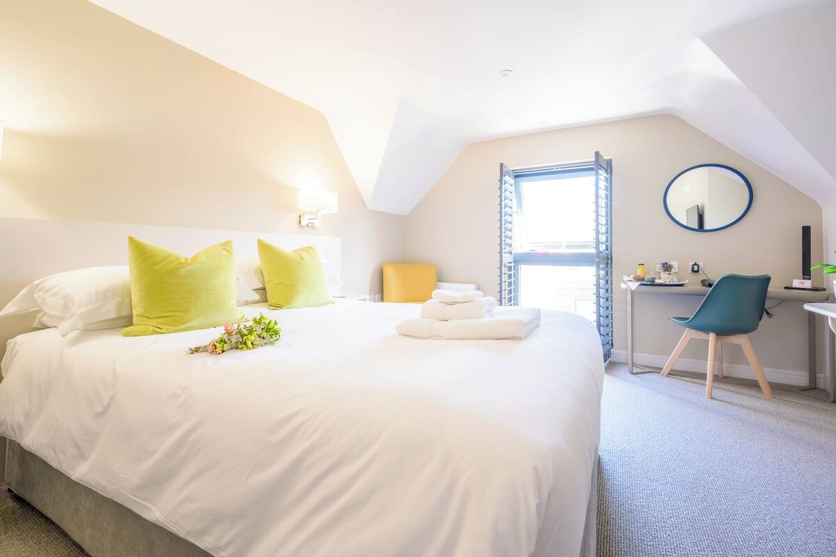 A hotel room with a large bed, a desk, a chair, a round mirror, and a window with blinds.