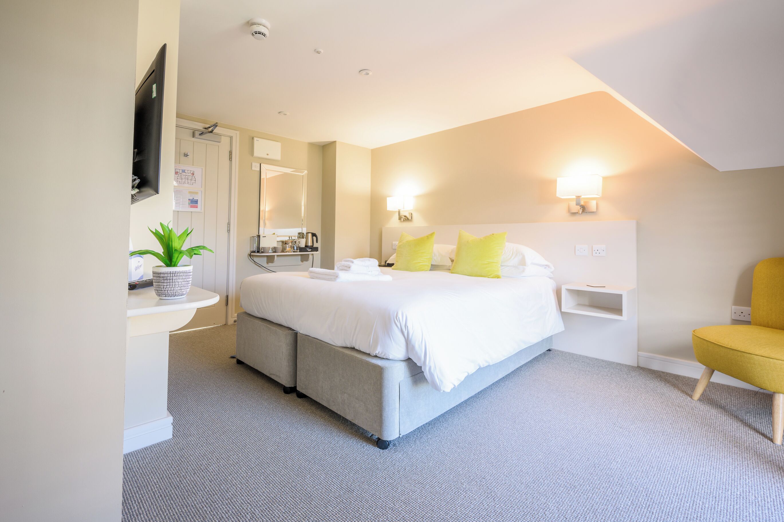 Een moderne slaapkamer met een groot bed, nachtkastjes, een flatscreen televisie, een kleine plant en een gele stoel.