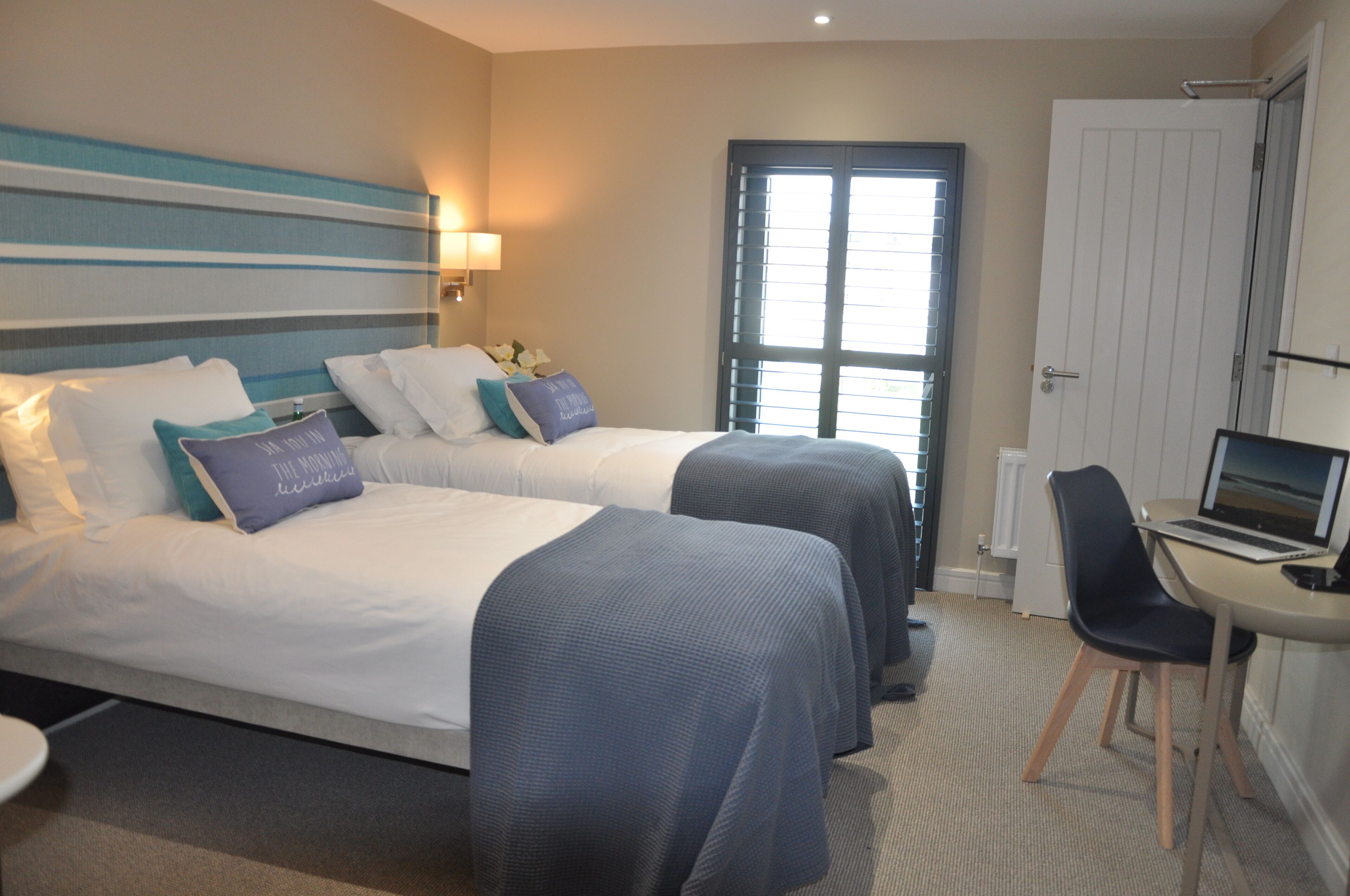 A hotel room with two beds, a desk with a laptop, a chair, and a window with blinds.