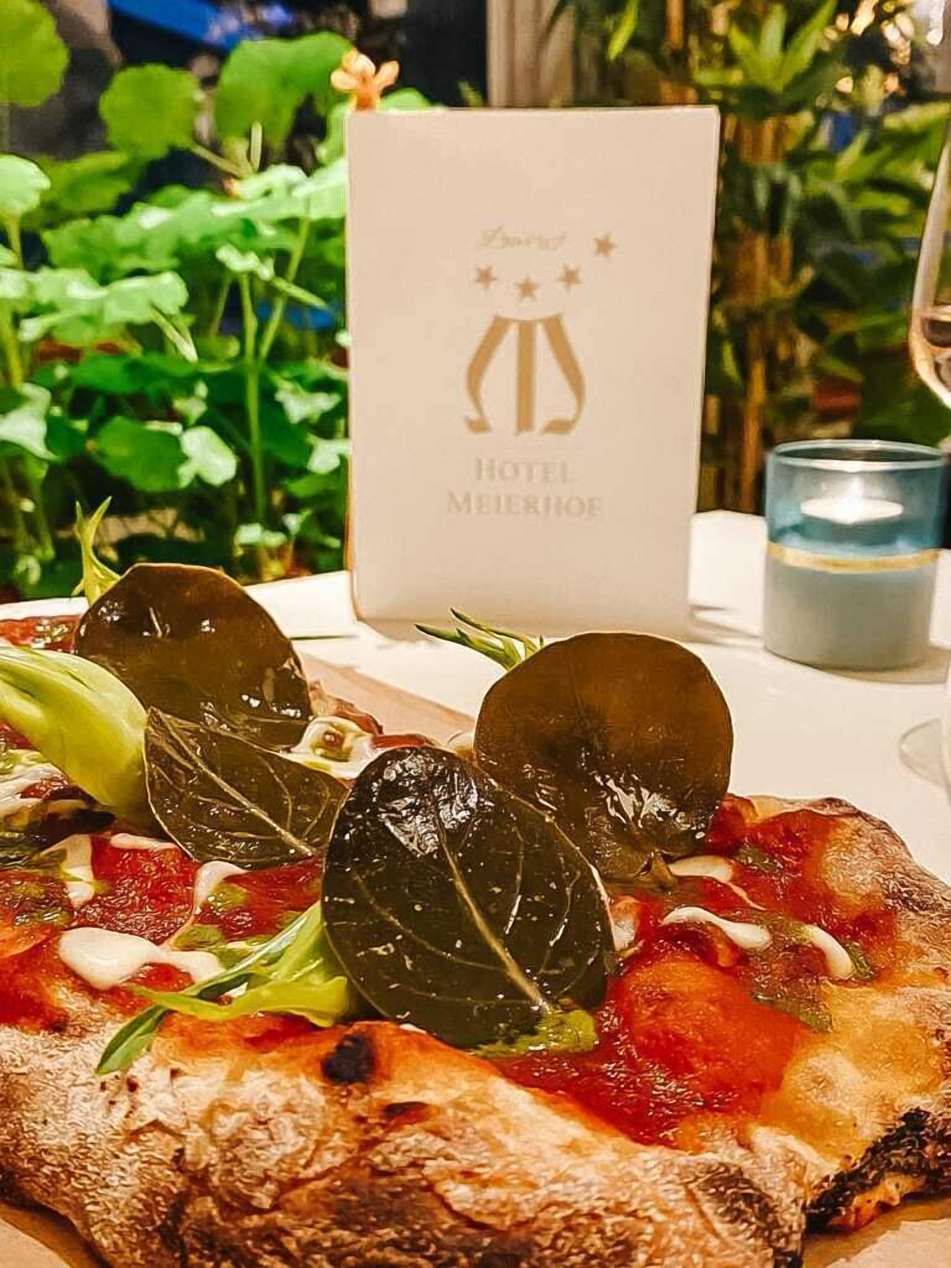 A pizza with toppings and a menu card labeled "HOTEL MEIERHOF" in the background.