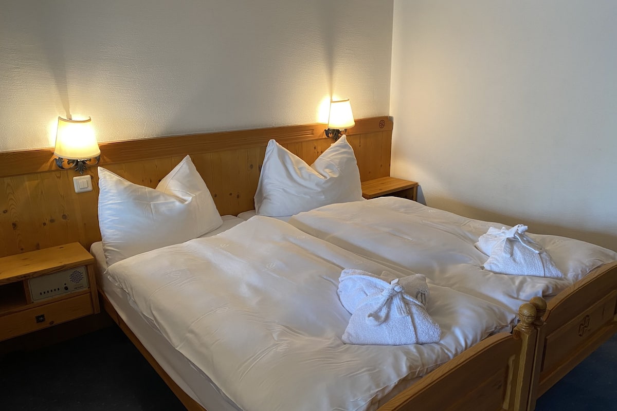 Una cama doble con sábanas blancas, dos lámparas de noche, un cabecero de madera y una mesita de noche con toma de corriente.