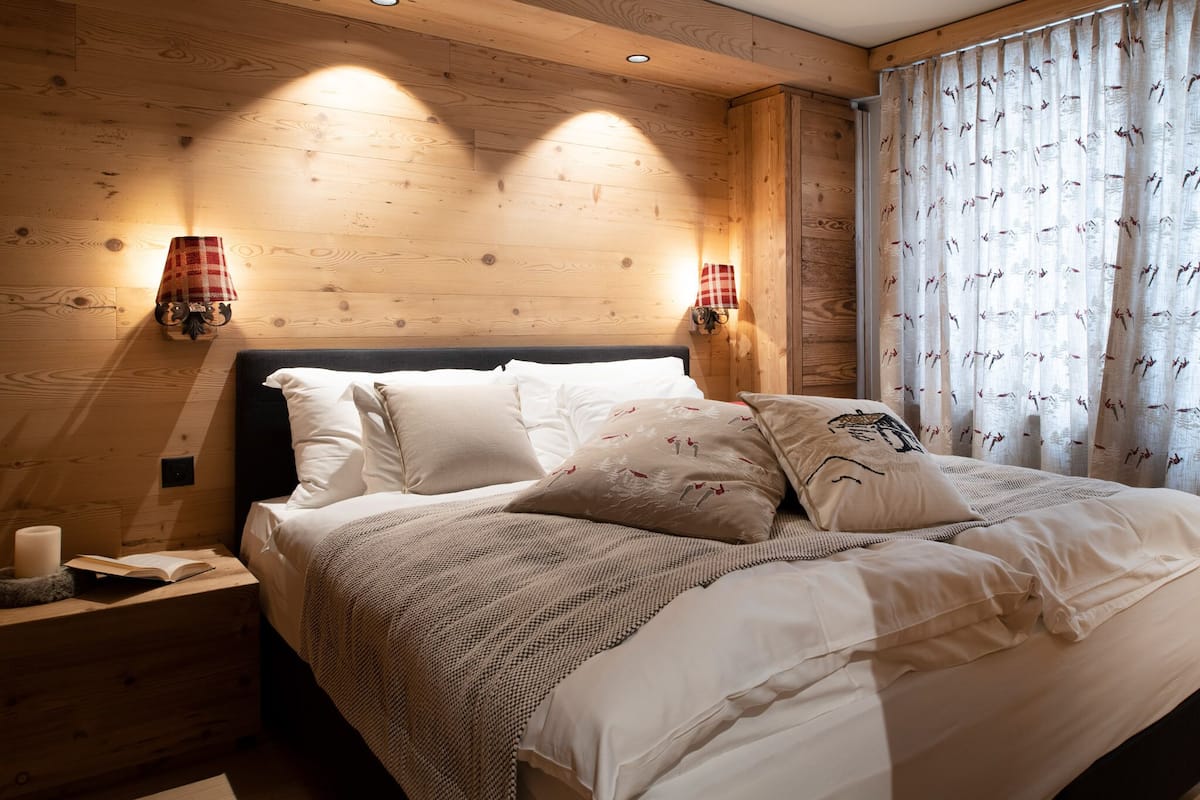 Una cama bien hecha con sábanas blancas y almohadones decorativos, cabecera de madera y dos lámparas fijadas a la pared.