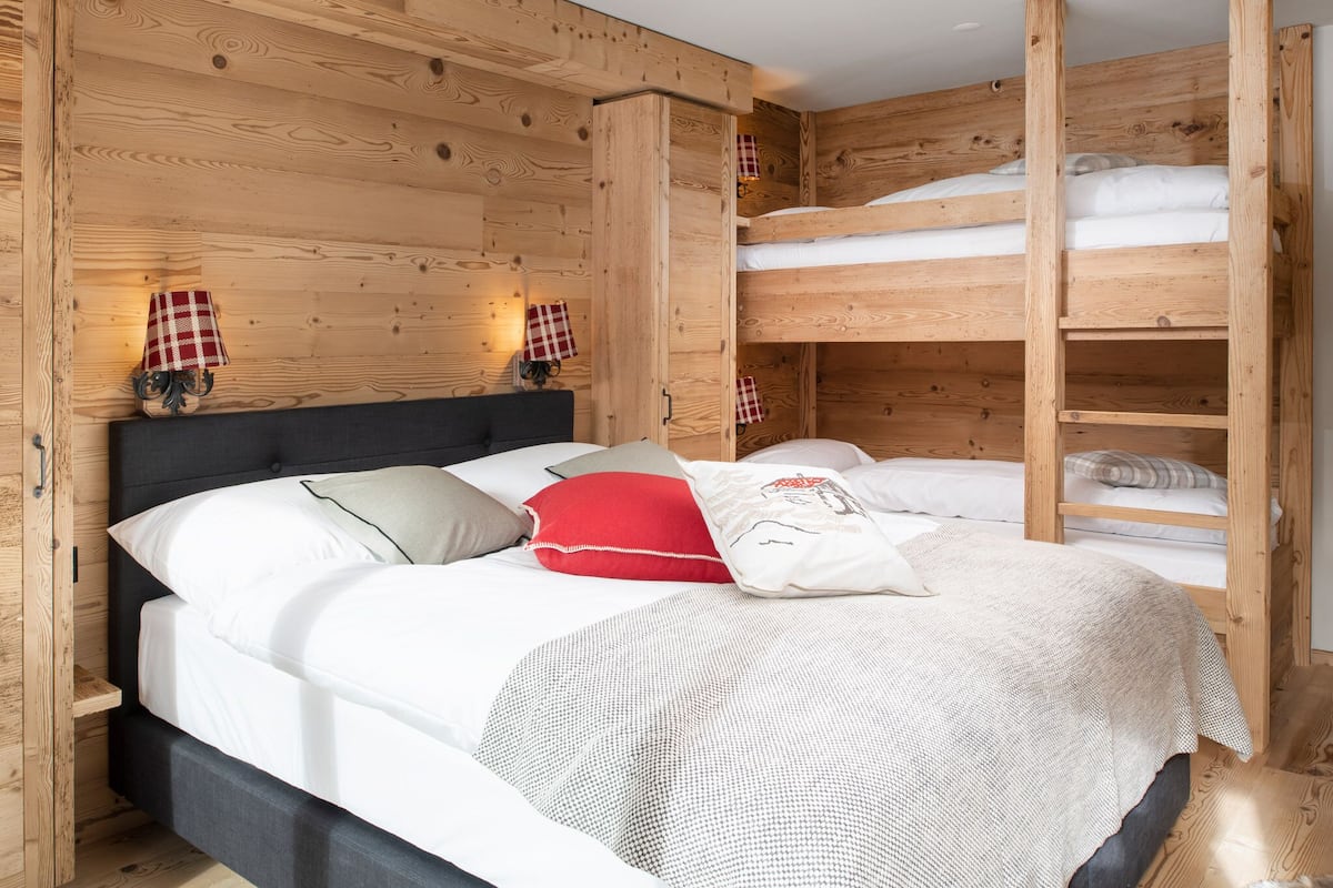 Una habitación tipo cabaña de madera con una cama litera, un cabecero oscuro y dos lámparas de pared con pantallas a cuadros rojos y blancos.