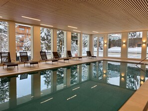 A modern indoor pool with clear glass walls, wooden ceiling, and multiple lounge chairs. - Residenz by Meierhof (Davos)