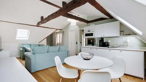 Un appartement de style « loft » avec des poutres apparentes en bois, une kitchenette, un coin repas avec une table ronde et des chaises blanches, et un canapé bleu clair.