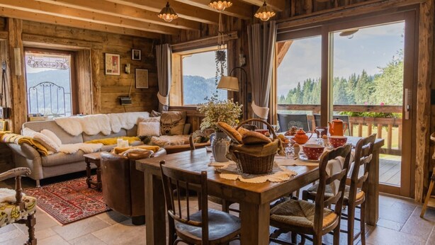 Interior acogedor de una cabaña de madera con comedor, sofá y ventanas amplias que ofrecen vistas al exterior.