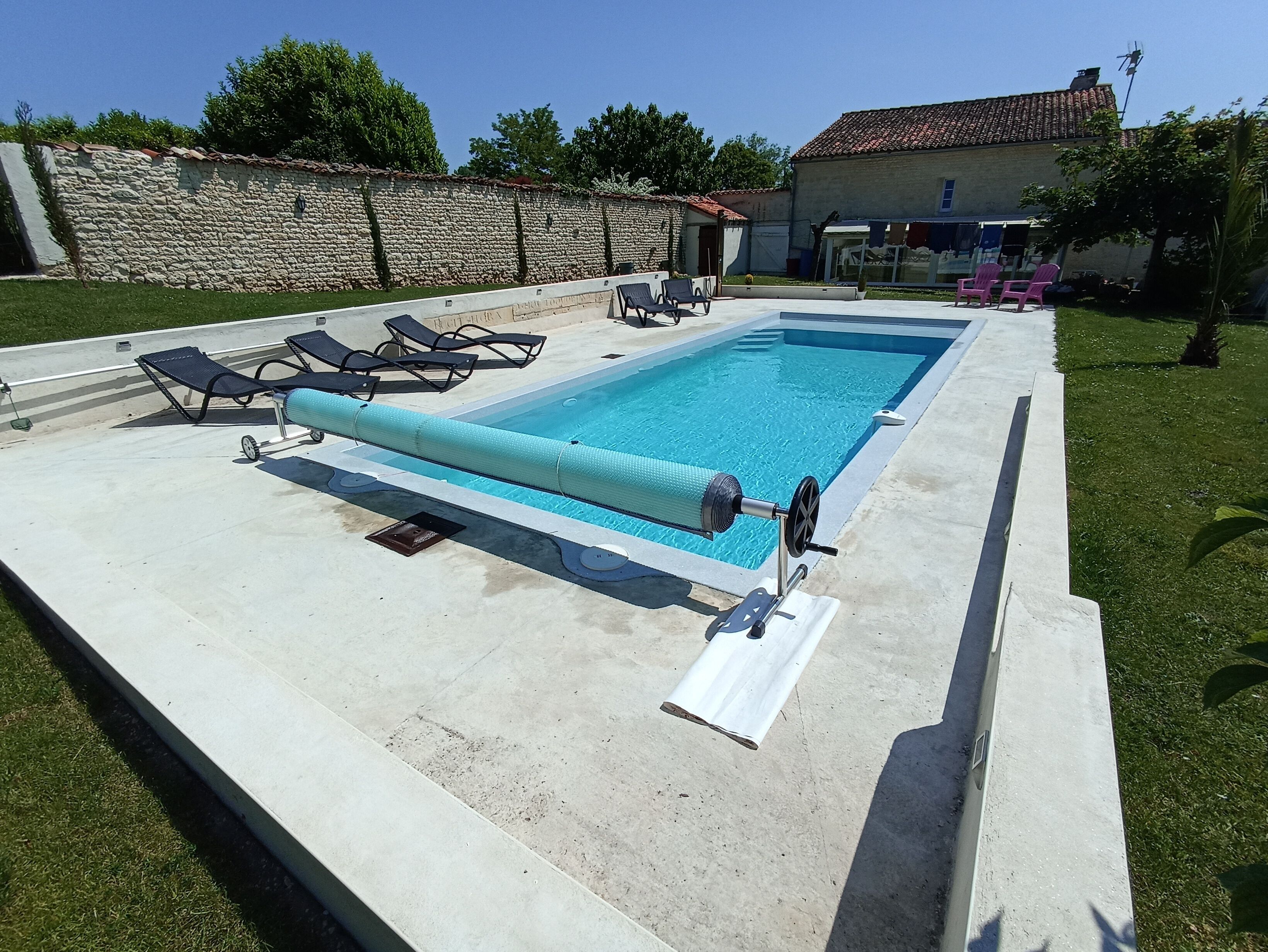 Una piscina rectangular con una cubierta, rodeada por una solana de hormigón. Hay sillas de descanso y una pequeña mesa cerca de la piscina.