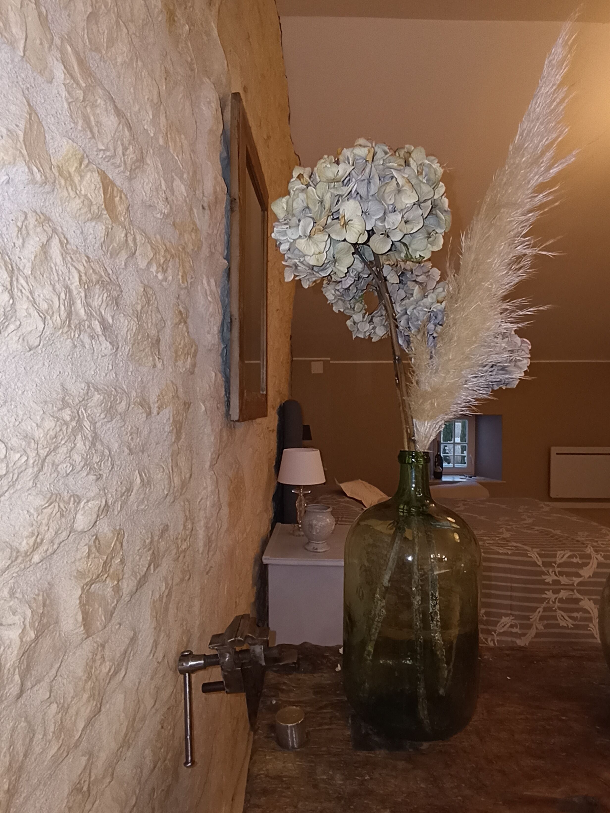 A room with a textured stone wall, a wooden bed, a bedside table with a lamp, and a vase with flowers and feathers.