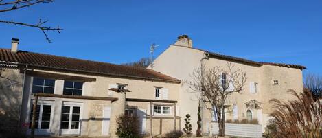 Un bâtiment en pierre de deux étages, doté d’un toit recouvert de tuiles, d’une entrée principale avec une porte et d’un petit jardin agrémenté d’un chemin menant à l’entrée.