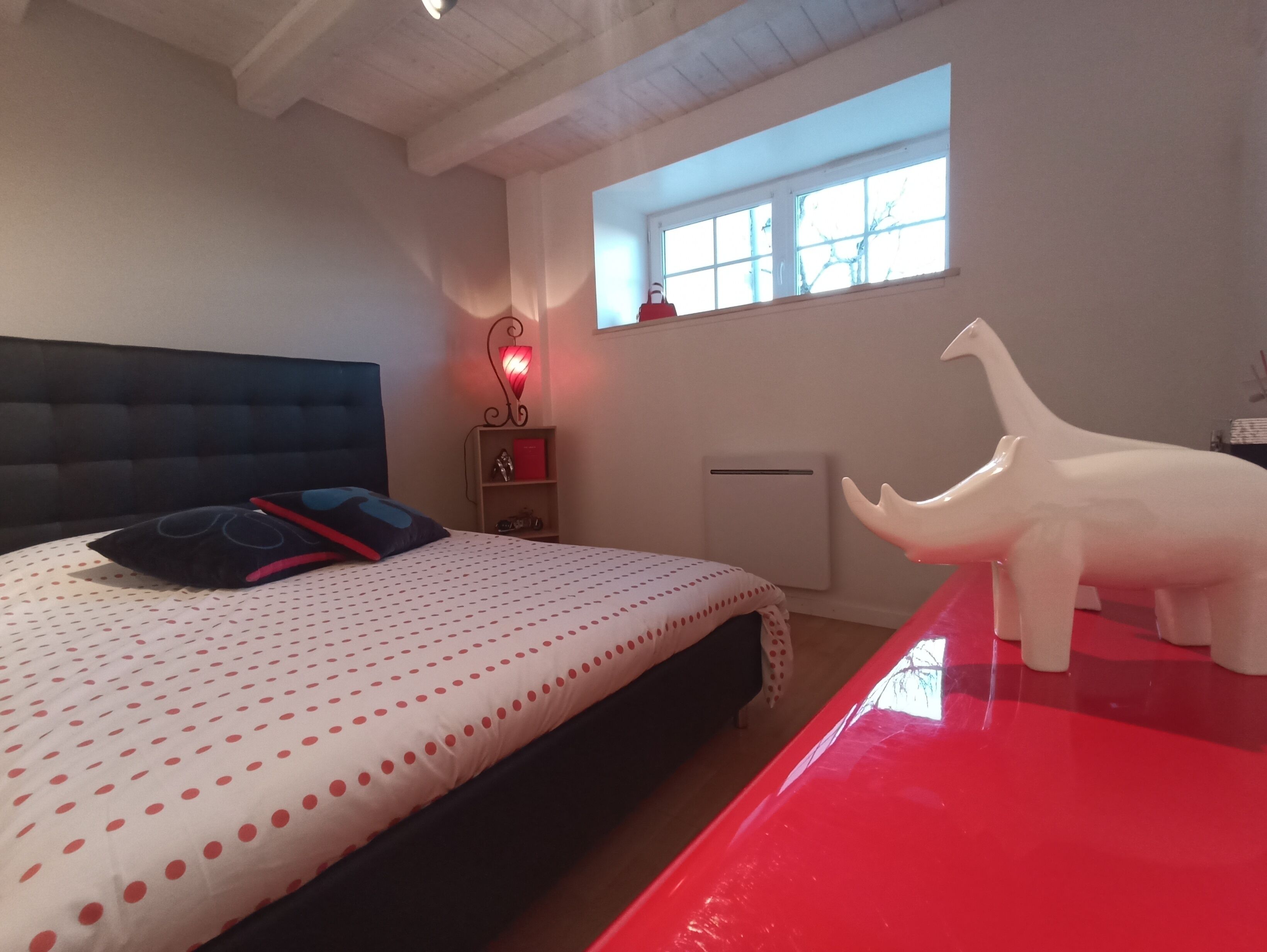 Ein Schlafzimmer mit einem Bett, einem roten Tisch, einer weißen Dinosaurierfigur, einer roten Lampe und einem Fenster.