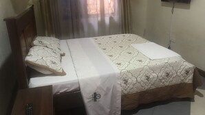 Ein ordentlich bezogenes Bett mit einem gemusterten Überwurf, ein Nachttisch mit Fernbedienung und ein an der Wand montierter Fernseher.