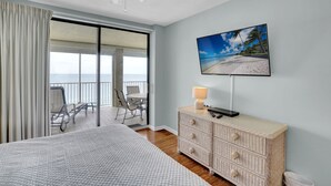 A hotel room with a bed, a television, a wicker dresser, and a balcony with a view of the beach. - White Caps 1506 by Liquid Life (Orange Beach)