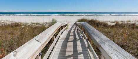 Ein Holzsteg führt zu einem Strand mit weißem Sand und im Hintergrund schwellenden Ozeanwellen.