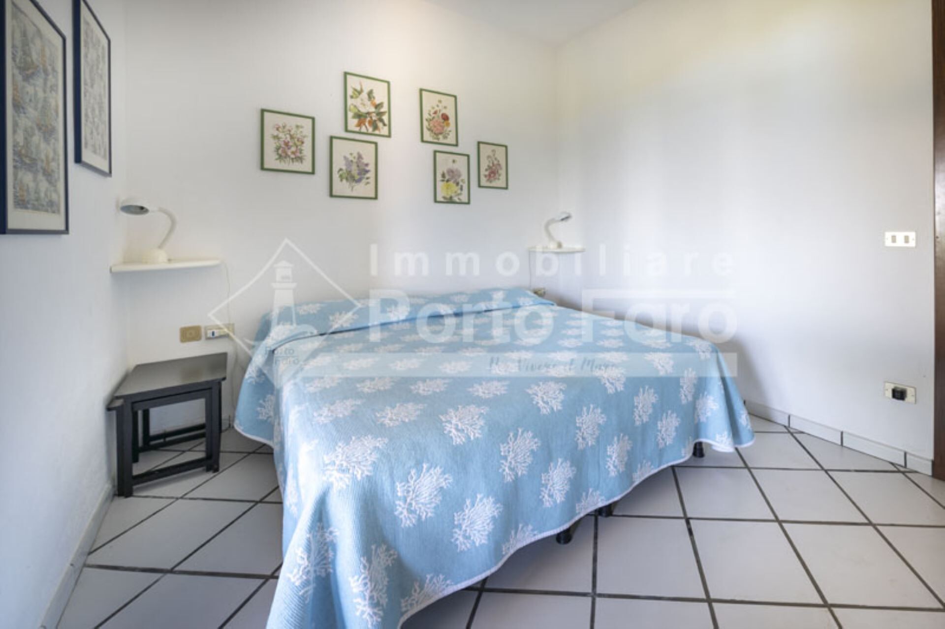 Ein Schlafzimmer mit einem Bett, das mit einer blau-blumigen Tagesdecke bezogen ist, einem Nachttisch mit einer Lampe und vier gerahmten Blumenbildern an der Wand.