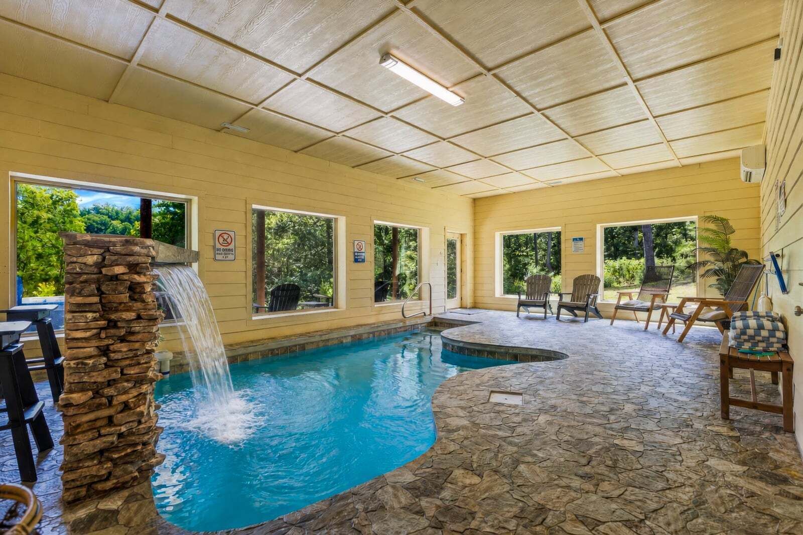 An indoor swimming pool with a stone waterfall feature, surrounded by stone flooring, and furnished with outdoor chairs and tables.
