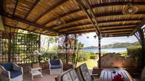 A covered outdoor seating area with wicker furniture, a wooden table, and a view of a lake and hills. - 13A Appartamento - 13A PORTO MANNU (Palau)