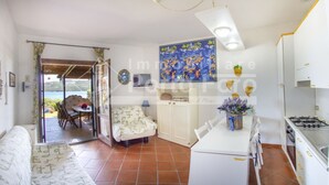 A cozy living room with a sofa, a dining table, and a kitchen area with a stove. There is a view of the outdoors through a glass door. - 13A Appartamento - 13A PORTO MANNU (Palau)