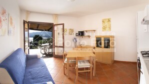 A compact living space with a sofa, a dining table with chairs, a kitchen area with a stove, and a sliding glass door leading to an outdoor patio with a view of mountains. - 15B Appartamento - 15B PORTO MANNU (Palau)