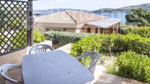A covered patio with a table and chairs, a view of a house with a tiled roof, and a fenced garden area. - 15B Appartamento - 15B PORTO MANNU (Palau)