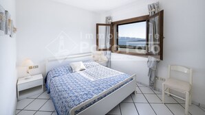 A bedroom with a bed, a chair, a window with curtains, and a nightstand with a lamp. - 16C Appartamento - 16C PORTO MANNU (Palau)