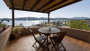 A covered patio with a round table and chairs, overlooking a lake and distant mountains. - 16C Appartamento - 16C PORTO MANNU (Palau)