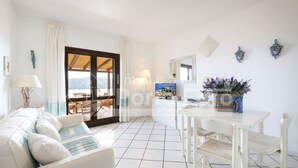 A bright living room with a sofa, a dining table, and chairs. There is a view through a glass door to an outdoor area with a table and chairs. - 16C Appartamento - 16C PORTO MANNU (Palau)