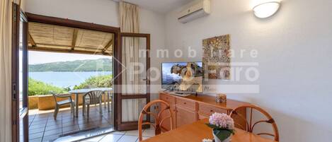 A room with a wooden dining table, a TV, and a view of the sea through a glass door.