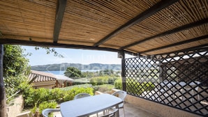 A covered outdoor seating area with a table and chairs, overlooking a scenic landscape with hills and a body of water. - 16B Appartamento - 16B PORTO MANNU (Palau)