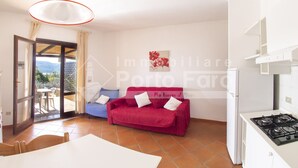 A cozy living room with a red sofa, a white table, and a kitchen area with a stove. - 16B Appartamento - 16B PORTO MANNU (Palau)