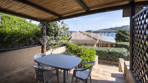 A covered patio with a table and chairs, a view of a house with a tiled roof, and a sign indicating a port. - 16B Appartamento - 16B PORTO MANNU (Palau)
