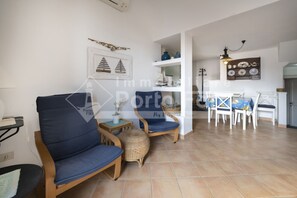 Un salon avec un canapé bleu, un fauteuil en osier et une table à manger avec des chaises. On y trouve une étagère avec des objets décoratifs et une lampe fixée au mur.