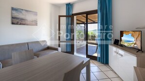 A modern living room with a sofa, a dining table, and a TV. There is a sliding glass door leading to an outdoor area. - 2B Appartamento - 2B PORTO MANNU (Palau)
