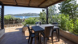 A covered patio with a table and chairs, a stone wall, and a view of the sea and hills. - 2B Appartamento - 2B PORTO MANNU (Palau)