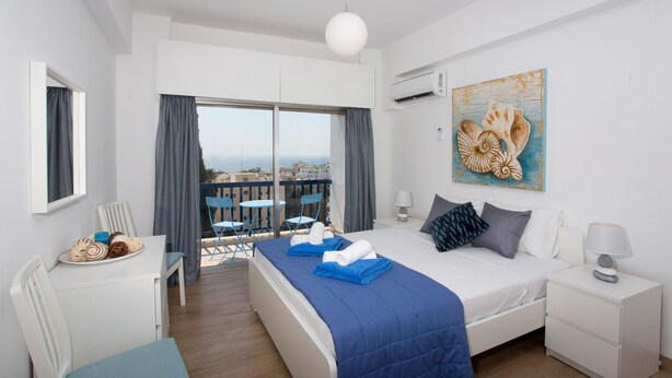 Una camera d'albergo con un letto, una scrivania con un piatto di pasticcini, un balcone con sedie e un quadro raffigurante conchiglie.