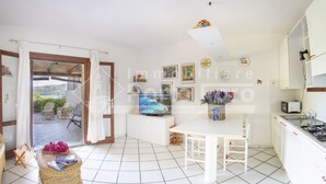 A bright kitchen with white cabinets, a dining table, and a TV. There is a view of the outdoors through an open door. - 20E Appartamento - 20E Porto Mannu (Palau)