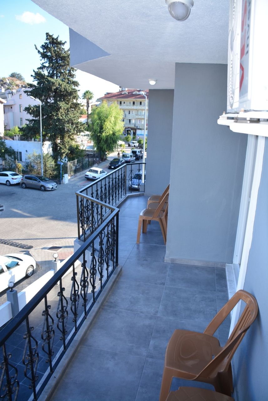 A balcony with a metal railing, two plastic chairs, and a view of a street with parked cars and buildings in the background.