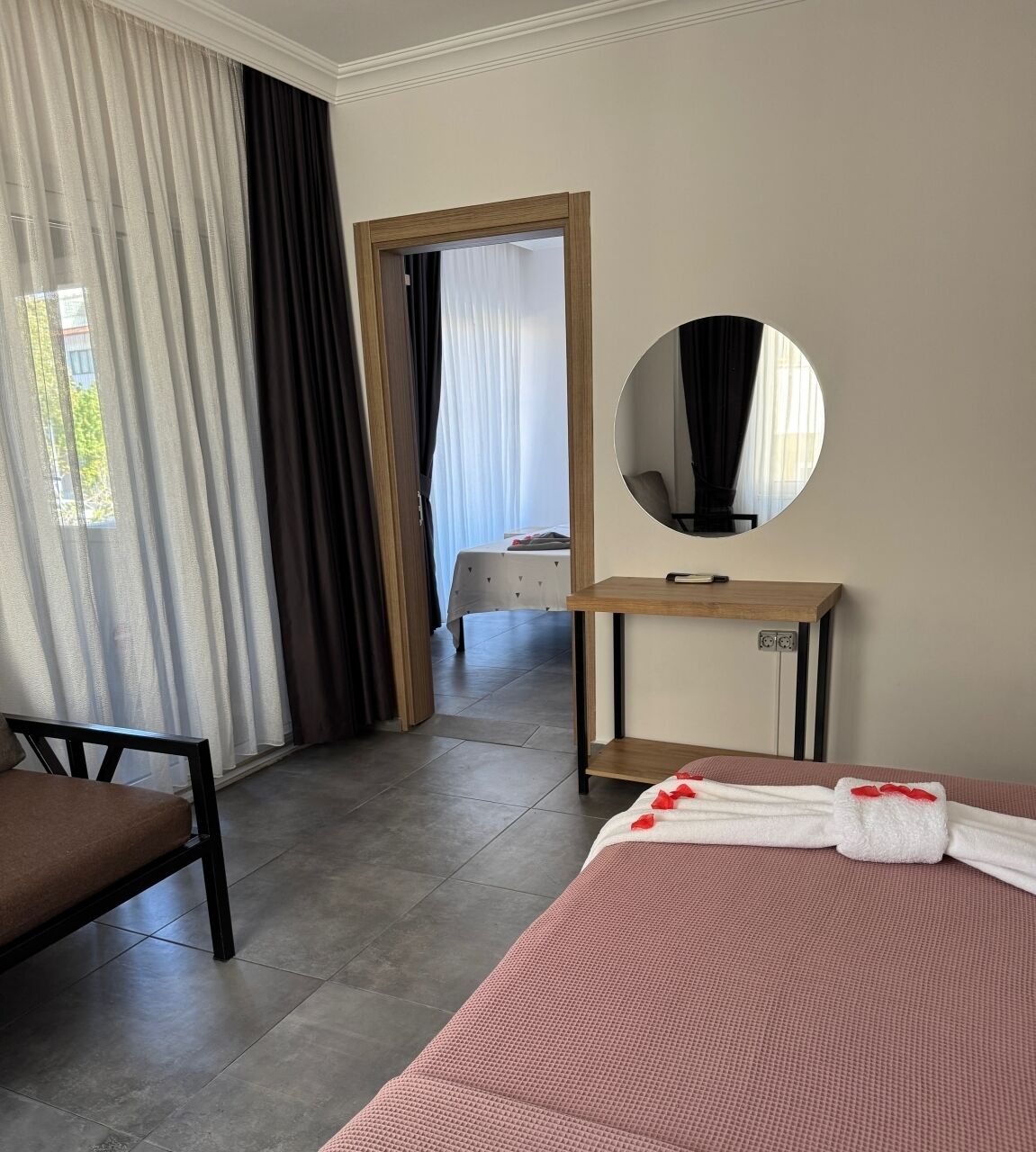 Una habitación de hotel con cama, una silla, una mesita de noche, un espejo y vista a otra habitación por una puerta.