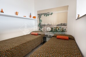 A room with two single beds, each with a patterned bedspread and orange pillows. A large painting adorns the wall behind the beds. - 9FARO - 9F PORTO FARO (Palau)