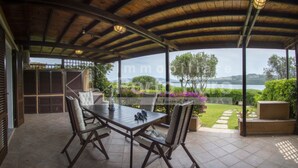 A covered patio with a table and chairs, a view of a lake, and a garden with a pathway. - 7A Appartamento - 7A PORTO MANNU (Palau)