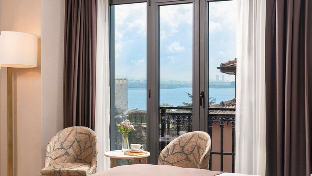 A hotel room with a bed, two chairs, a small table with a vase and a cup, and a view of a cityscape through large windows.