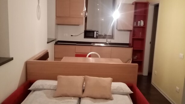 A bedroom with a bed, a bedside table, a red chair, a small desk, a mirror, and a kitchenette with a microwave. - ex scuola ristrutturata di recente (PAULARO)