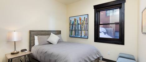 A bedroom with a bed, bedside table, lamp, and a window with a view of a building.