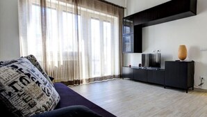 A modern living room with a dark sofa, a flat-screen TV on a black stand, and a large window with sheer curtains. - Appartamento Ariel   La Perla del Mare di Polignano (Polignano a Mare)