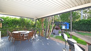 Un espace extérieur abrité, doté d’une terrasse en bois, d’une table ronde avec des chaises et d’un barbecue.