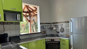 Une cuisine avec des meubles de couleur verte, un four Bosch, un réfrigérateur en acier inoxydable et une fenêtre donnant sur des arbres et une structure en bois.